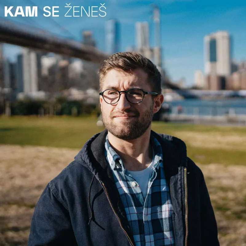 Muž s okuliarmi vo flanelovej košeli stojí v slnečnom parku s mestskou krajinou v pozadí. V ľavom hornom rohu je nápis „KAM SE ŽENEŠ“.