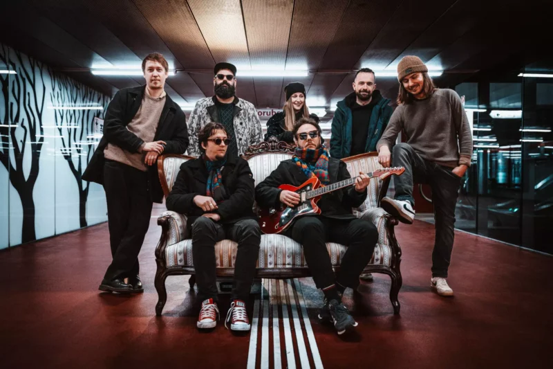 Sedem ľudí pózuje v modernej chodbe s nástennými maľbami stromov. Dvaja sedia na vintage pohovke, jeden drží gitaru, všetci vyžarujú uvoľnenú, kreatívnu atmosféru.