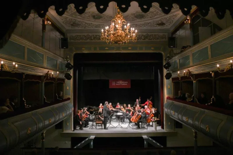 Komorný orchester vystupuje na javisku v zdobenom divadle. Hudobníci sú formálne oblečení a nad nimi visí luster, ktorý vytvára elegantnú atmosféru.