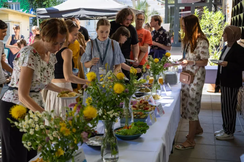 Ľudia sa zhromažďujú okolo bufetového stola s farebnými jedlami a kvetinovými dekoráciami na podujatí pod holým nebom. Ľahká, neformálna atmosféra za slnečného dňa.