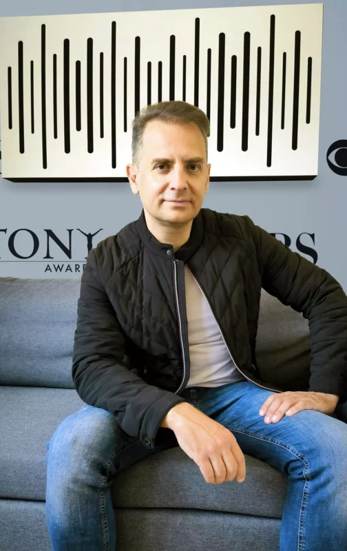 Na sivom gauči sedí osoba v čiernej bunde a džínsoch s pokojným výrazom. Za ňou je stena so zvislými čiernymi čiarami na bielom paneli.