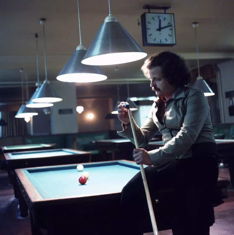 Muž s kučeravými vlasmi sedí na biliardovom stole a kreslí kriedou tágo. Stropné svetlá osvetľujú scénu a vytvárajú pokojnú a sústredenú atmosféru.