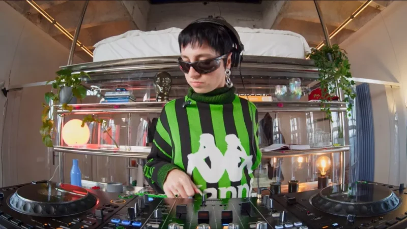 DJ v zeleno-čiernom svetri so slúchadlami na ušiach mixuje hudbu na gramofóne. Prostredie je moderné a štýlové, s rastlinami a jemným osvetlením.