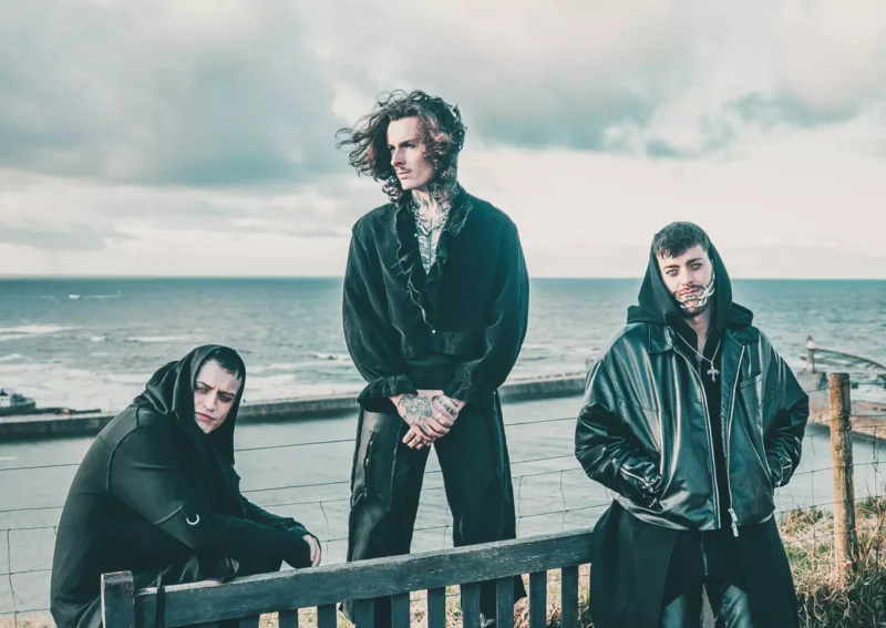 Traja ľudia v tmavom, módnom oblečení pózujú pri oceáne. Scéna je melancholická, so zamračenou oblohou a vzdialeným, pokojným horizontom.