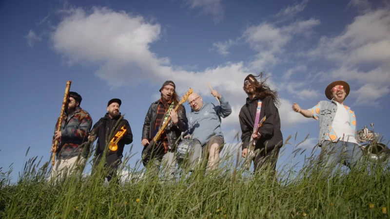 Živá kapela hrajúca na slnečnom poli, každý člen s inými nástrojmi ako flauta a gitara, na pozadí modrej oblohy a oblakov.