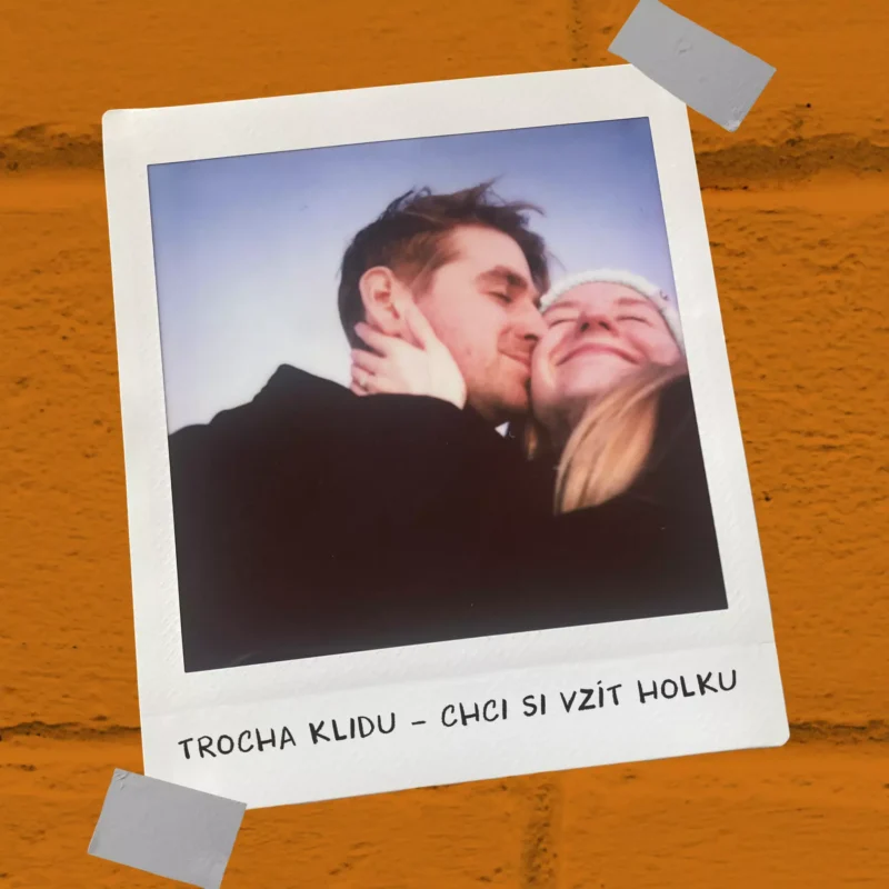 Pár sa objíma a šťastne sa usmieva na polaroidovej fotografii pri oranžovej tehlovej stene. Fotografia nalepená na stene vyžaruje teplo a radosť.
