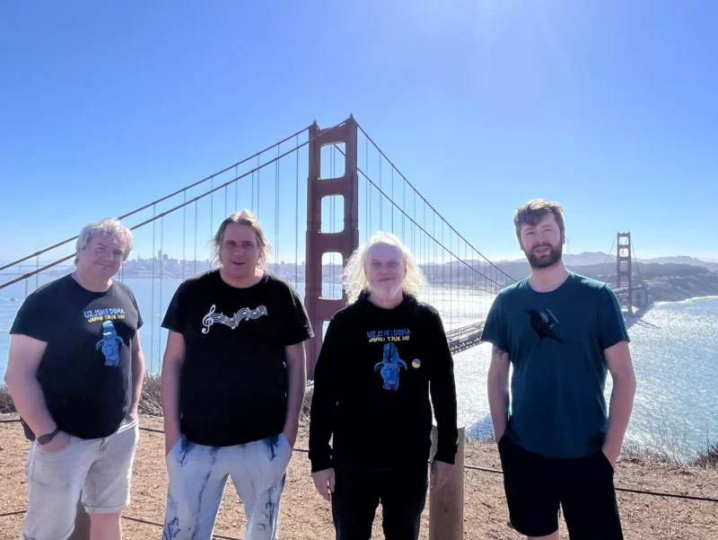 Štyria ľudia stoja s úsmevom pred slnkom zaliatym mostom Golden Gate. Jasná obloha a trblietavá voda vytvárajú veselú a živú atmosféru.