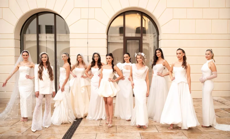 Skupina žien v elegantných bielych šatách stojí sebavedomo pred klenutými oknami na kamennej terase a vyžaruje z nich pôvab a štýl.
