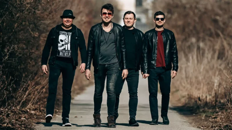 Štyria muži v čiernych kožených bundách sebavedomo kráčajú po vidieckom chodníku, po oboch stranách ktorého sú suché kríky. Nálada je ležérna a uvoľnená.