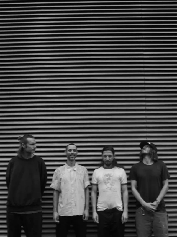 Štyria muži stoja pred pruhovanou stenou, každý s jedinečnou pózou. Čiernobiela fotografia vyvoláva ležérnu a uvoľnenú náladu.