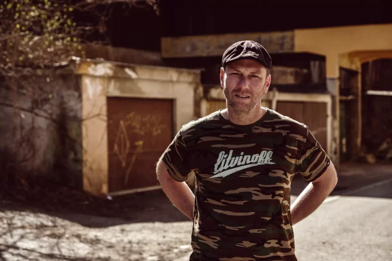 Muž v maskáčovom tričku a čiapke stojí vonku pred starou budovou s graffiti. Má uvoľnený výraz a scéna pôsobí ležérne a rustikálne.