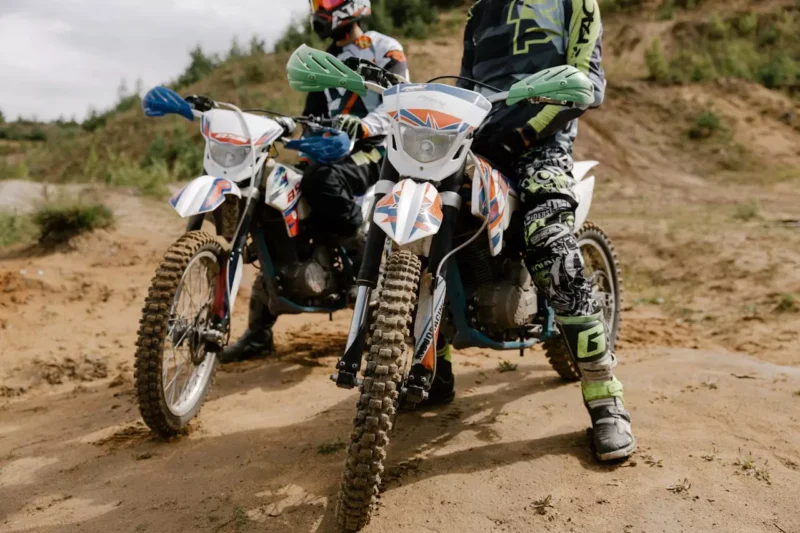 Dvaja ľudia v motokrosovom oblečení sedia na terénnych motorkách s hrboľatými pneumatikami, vyvážení na piesočnatej ceste obklopenej zeleňou, a vyjadrujú vzrušenie a dobrodružstvo.