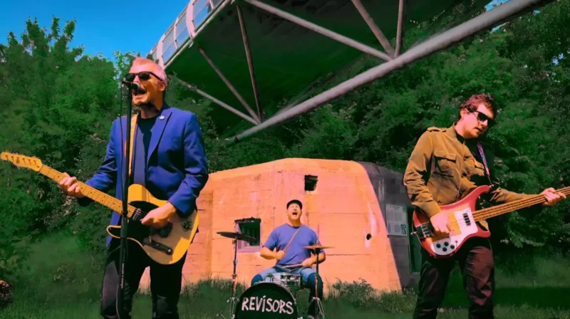 Kapela energicky vystupuje vonku. Hlavný spevák v modrej bunde hrá na elektrickú gitaru, po boku bubeníka a basgitaristu. Jasná, živá scéna.