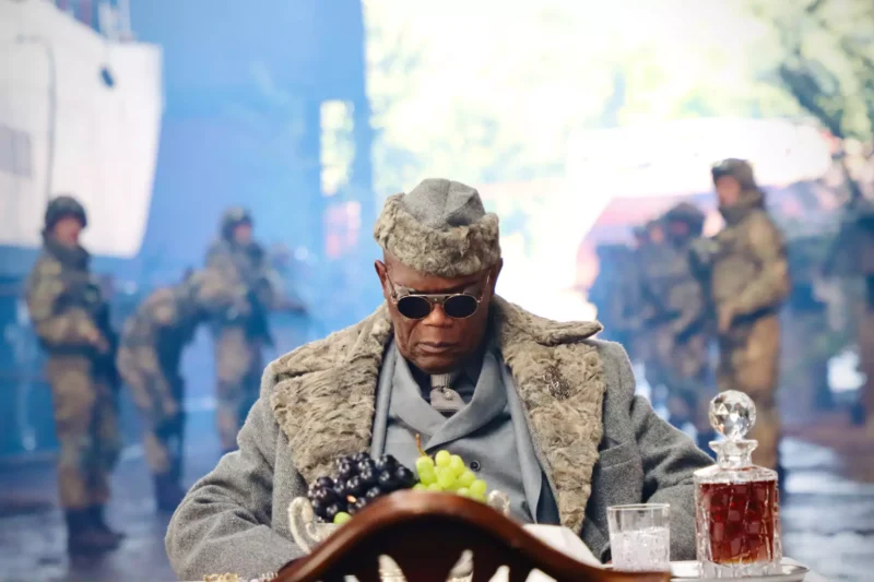 Muž v kožuchu a slnečných okuliaroch sedí pri stole s hroznom a karafou. Rozmazané pozadie zobrazuje vojakov, čo vytvára napätú atmosféru.