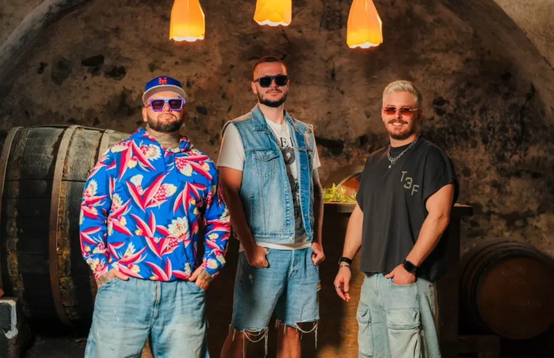 Traja muži so slnečnými okuliarmi, oblečení v ležérnom oblečení, sebavedomo stoja v slabo osvetlenej pivnici so stenami lemovanými sudmi a oranžovými závesnými lampami nad nimi.