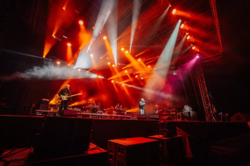 Koncertné pódium so žiarivými oranžovými a bielymi svetlami svietiacimi nadol. Kapela energicky vystupuje pred veľkým publikom za hranicami dohľadu. Elektrizujúca atmosféra.