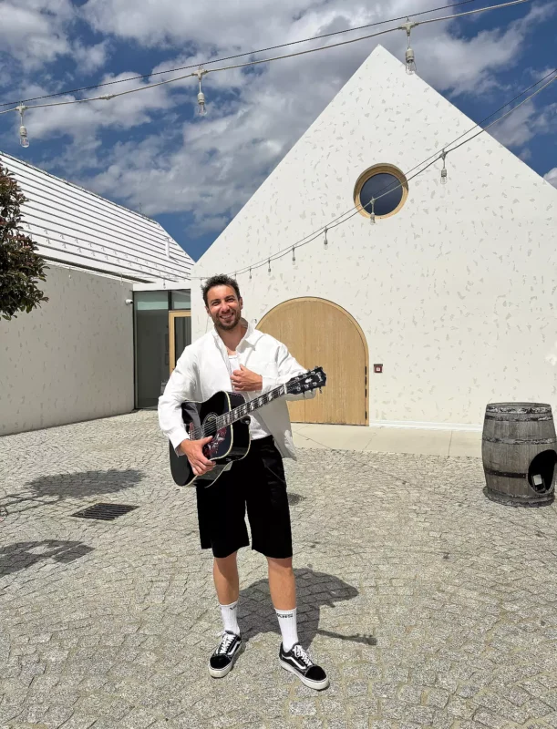 Usmievavý muž stojí na dláždenom dvore a hrá na gitare. Má na sebe bielu košeľu a čierne šortky. V pozadí je moderná budova a jasná obloha.