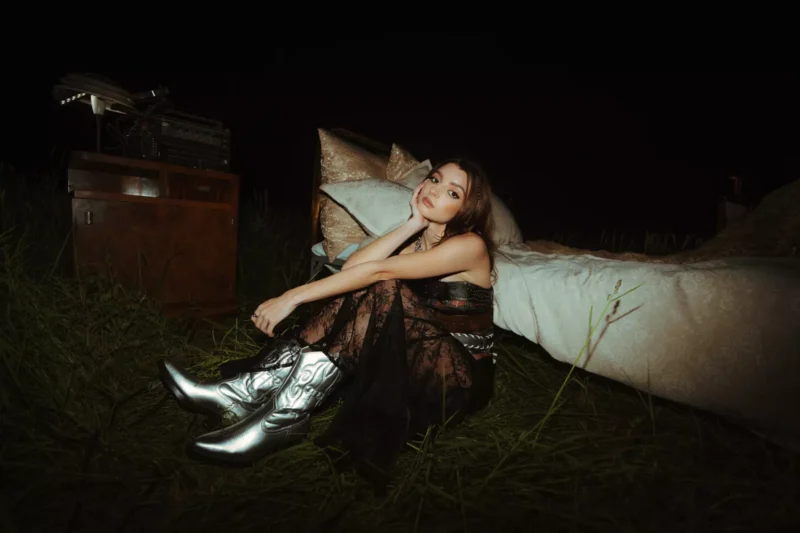 Žena v čiernych čipkovaných šatách a strieborných čižmách zamyslene sedí v noci vo vysokej tráve, opretá o posteľ. Neďaleko na drevenej skrinke je starožitné rádio.