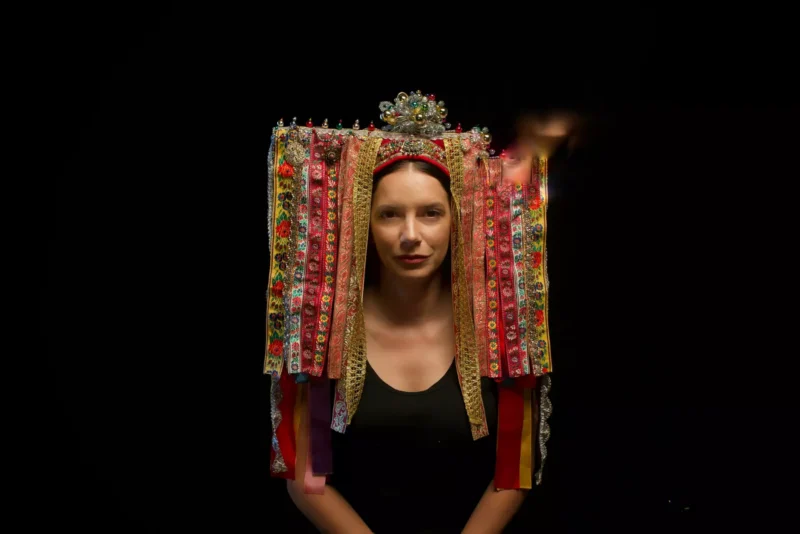 Žena v čiernom tope má na sebe tradičnú, zdobenú pokrývku hlavy s farebnou kvetinovou výšivkou a stuhami na tmavom pozadí, ktorá vyjadruje kultúrne bohatstvo.