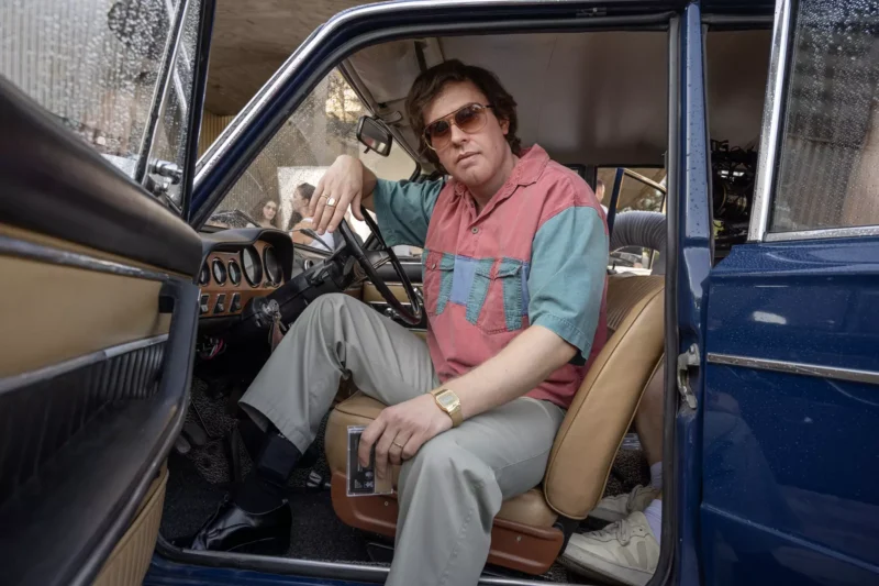 Muž v starožitnom aute so slnečnými okuliarmi a farebnou košeľou drží kazetu v ruke. Otvorené dvere odhaľujú daždivú scenériu vonku s uvoľnenou atmosférou.