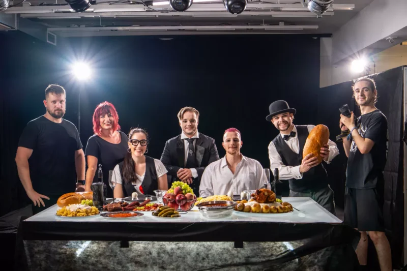 Skupina siedmich ľudí stojí za banketovým stolom prestretým rôznymi jedlami, usmieva sa pod jasnými štúdiovými svetlami a vytvára živú, slávnostnú atmosféru.