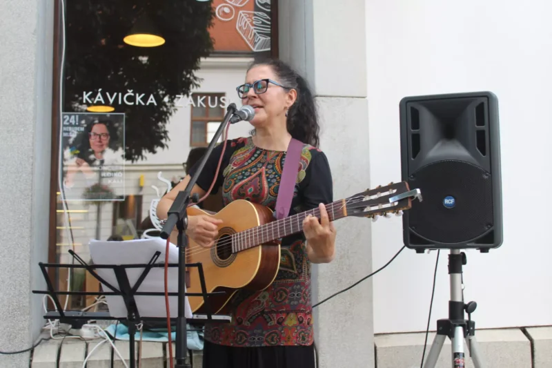 Žena spieva a hrá na gitare pred oknom kaviarne, má na sebe okuliare a vzorované šaty. Na stojanoch je mikrofón a notový záznam a neďaleko je reproduktor.
