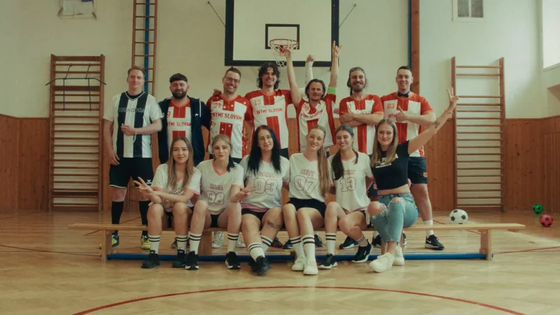 Radostná skupinka mladých dospelých, niektorí v športových dresoch a pruhovaných ponožkách, pózuje v telocvični s basketbalovým košom a farebnými futbalovými loptami a vyžaruje z nich tímového ducha.