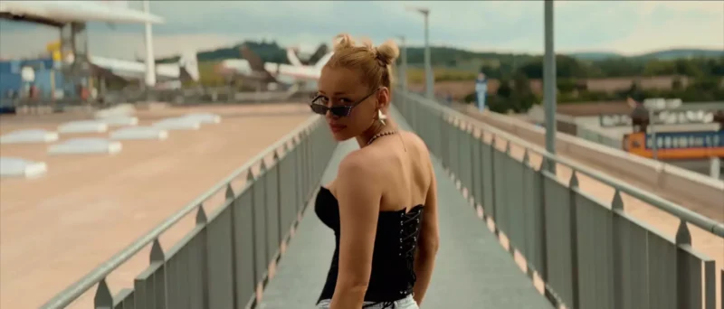 Osoba s dvoma drdolmi, slnečnými okuliarmi a čiernym topoľom sa pozerá späť na most. V pozadí je rozmazaná priemyselná zóna.