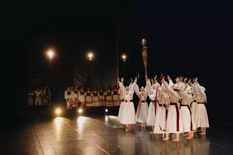 Skupina účinkujúcich v bielych tradičných krojoch s červenými šerpami energicky tancuje na pódiu; za nimi stojí rad hudobníkov v podobnom oblečení. Scéna je príjemne osvetlená, čo vytvára atmosféru kultúrnej oslavy.