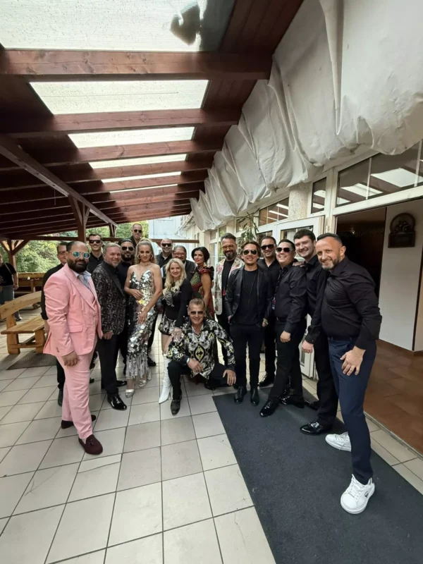 Skupina ľudí vo formálnom oblečení, vrátane ružového obleku a flitrovaných šiat, pózuje pod krytou terasou. Vyzerajú veselo a oslavne.