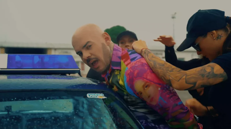 Plešatého muža v farebnej košeli tlačí k policajnému autu potetovaný policajt s prísnym výrazom. Nálada je napätá a dramatická.