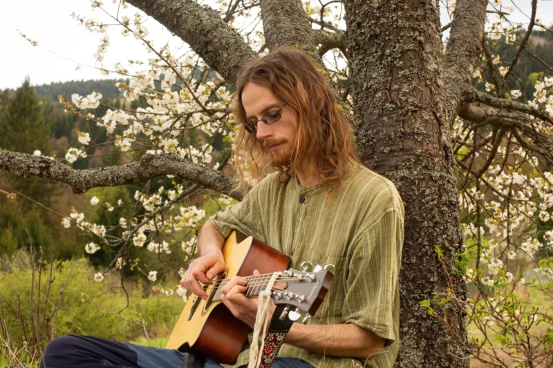 Muž s dlhými vlasmi a okuliarmi hrá na akustickú gitaru, zatiaľ čo sedí pod kvitnúcim stromom. Scéna vyvoláva pokojnú, rustikálnu atmosféru.