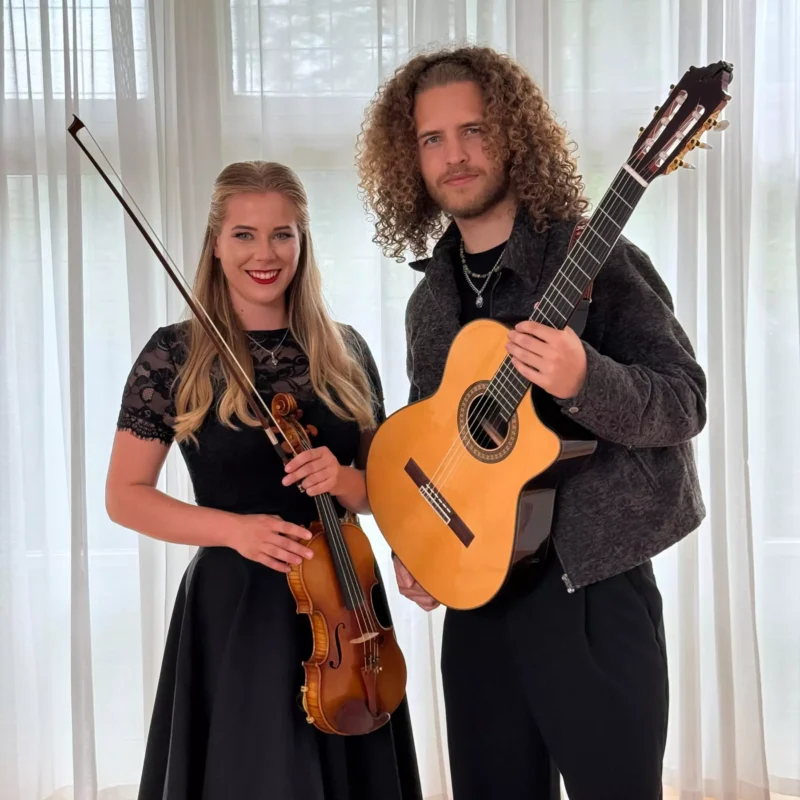 Žena a muž sa vrúcne usmievajú a držia v rukách husle a gitaru. Stoja pred bielymi závesmi, čo naznačuje formálne, no zároveň uvoľnené prostredie.