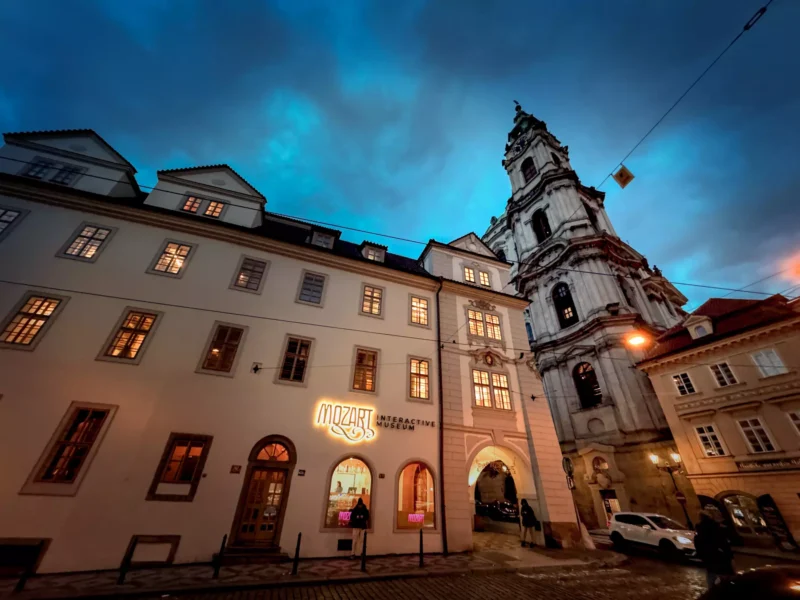 Historická európska ulica za súmraku so žiariacimi oknami. Na viditeľnom mieste je vystavené „Interaktívne múzeum Mozarta“. Dramatická obloha, dláždený chodník.