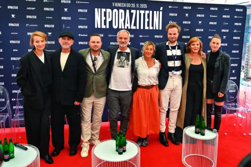 Skupina ôsmich ľudí stojí na červenom koberci na mediálnej akcii a vrúcne sa usmieva. Pozadie s tučným nápisom „Neporazitelní“ udáva profesionálny tón.