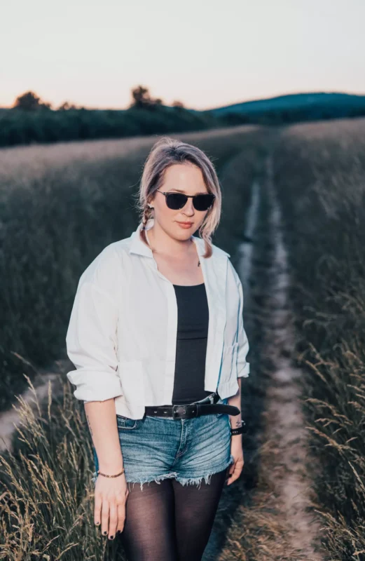 Žena so slnečnými okuliarmi stojí na trávnatom chodníku pri západe slnka, má na sebe bielu košeľu, čierny top a džínsové šortky. Scéna pôsobí pokojne a uvoľnene.