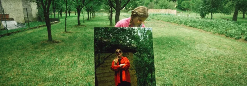 Žena sa pozerá do zrkadla na trávnatej lúke obklopenej stromami. Zrkadlo ju odráža v slnečných okuliaroch a červenom kabáte, čo vytvára surrealistický efekt.