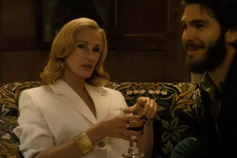 Žena v bielom obleku so zlatým náramkom sedí na vzorovanej pohovke s pohárom vína v ruke a rozpráva sa s bradatým mužom, čím vytvára intímnu a vrúcnu atmosféru.