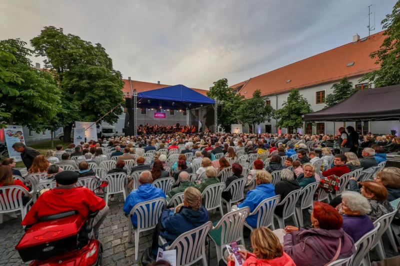 Vonkajšia koncertná scéna s veľkým sediacim publikom pred krytým pódiom. Podujatie lemujú stromy a atmosféra je živá a pútavá.
