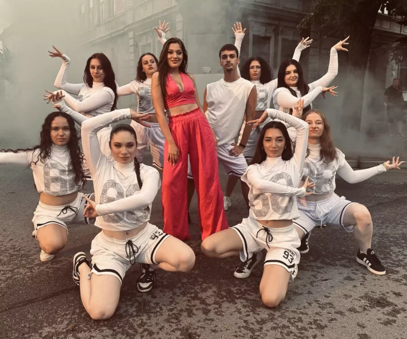 Skupina tanečníkov pózuje na ulici zahalenej v hmle. Ústredná postava má na sebe jasne ružový outfit, obklopená ďalšími v bielom športovom oblečení, vyžarujúca energiu a jednotu.