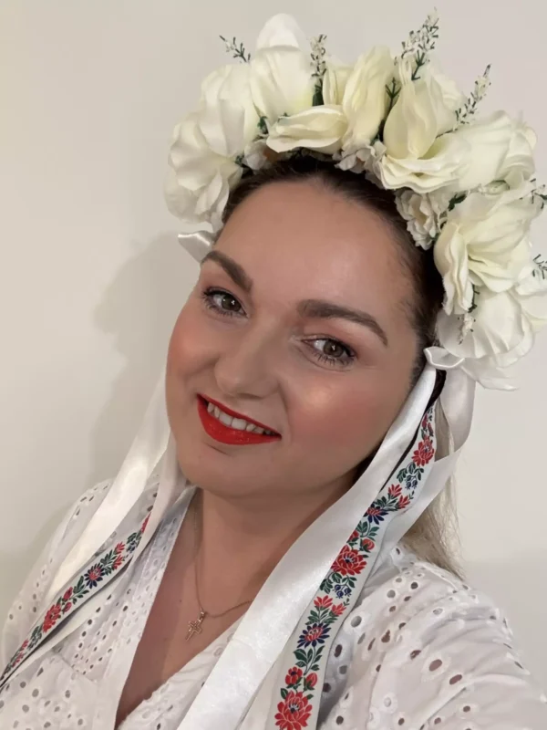 Usmievavá žena v bielej kvetinovej čelenke a tradičnom vyšívanom odeve s červenými a modrými vzormi, vyjadrujúca radosť a kultúrnu hrdosť.