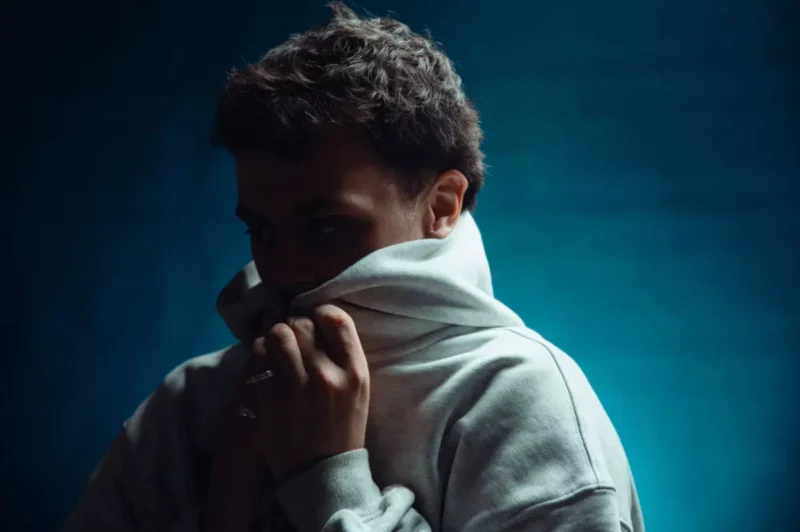 Osoba s krátkymi vlasmi si čiastočne zakrýva tvár sivou mikinou s kapucňou na pochmúrnom modrom pozadí, čo vyvoláva pocit introspekcie alebo rozjímania.