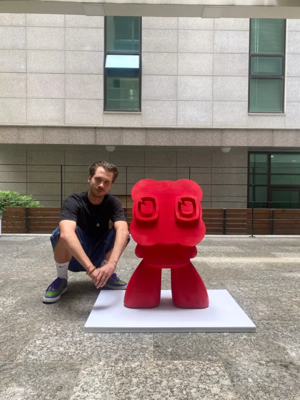 Muž sa krčí vedľa veľkej červenej abstraktnej sochy na bielom podstavci. Dej je mestský dvor so sivou dlažbou a modernou budovou v pozadí.