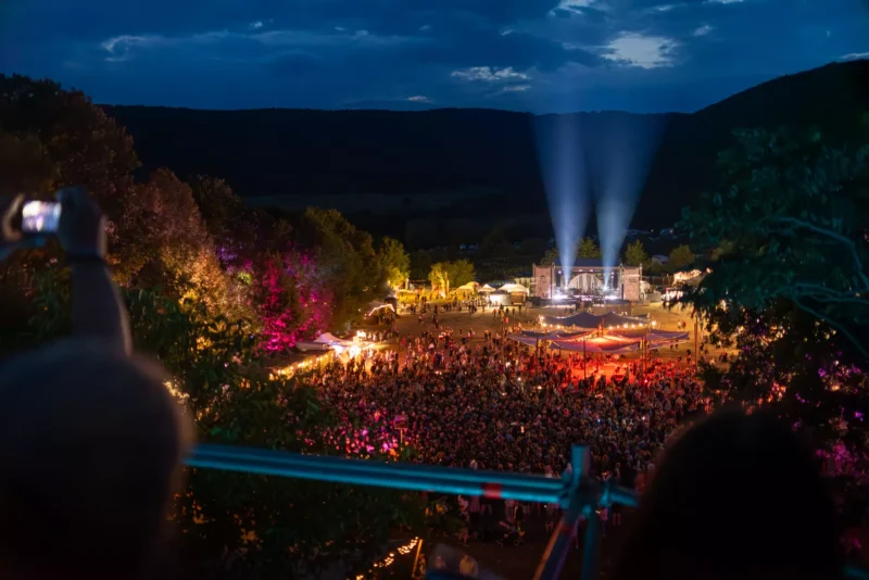 Večerný pohľad na hudobný festival pod holým nebom, kde je uprostred lúky pódiová scéna so svetelnými reflektormi smerujúcimi do neba. Okolo pódia sa zhromaždilo veľké množstvo ľudí, stromy a stánky navôkol sú osvetlené farebnými svetlami. Vpredu je rozmazaná silueta človeka s mobilom, ktorý si pravdepodobne fotí dianie na festivale.