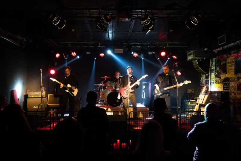 Na fotografii je štvorčlenná rocková kapela vystupujúca na klubovom pódiu s čiernym pozadím a efektovým osvetlením v odtieňoch modrej a červenej. V strede hrá bicista, vpredu sú gitarista, hlavný spevák s elektrickou gitarou a basgitarista, pričom každý má pri sebe mikrofón. Pred pódiom stoja diváci, priestor je ozdobený plagátmi na stenách a po okrajoch sú viditeľné hudobné aparáty a vybavenie.