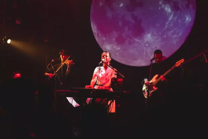 Na fotografii je koncertná scéna s klaviristkou a speváčkou, ktorú sprevádza huslista a basgitarista. V pozadí dominuje veľká projekcia mesiaca osvetľujúca priestor v jemných ružovo-fialových tónoch. Atmosféra je magická a snivá, publikum sleduje vystúpenie v prítmí.