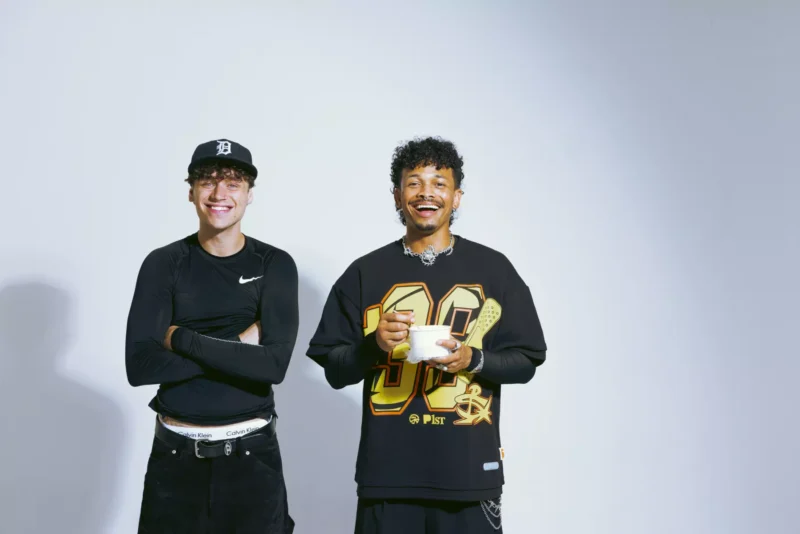 Dvaja muži sa usmievajú na svetlom pozadí. Jeden má na sebe čierny klobúk a rolák so skríženými rukami. Druhý má na sebe grafické tričko a v ruke drží pohár. Ležérna a veselá nálada.