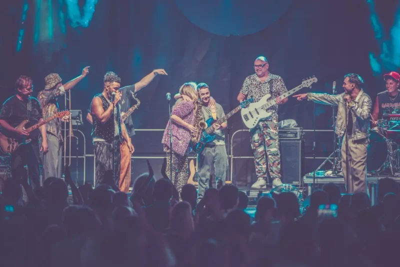 Na fotografii je veľká hudobná skupina vystupujúca na pódiu pred početným publikom. Členovia kapely majú pestrofarebné, extravagantné kostýmy a hrajú na rôzne nástroje – elektrické gitary, basgitaru, saxofón či klávesy. V strede sa objímajú dvaja hudobníci, ostatní gestikulujú alebo spievajú do mikrofónov, atmosféra je energická, svetlá sú modré a pódiová scéna pôsobí živo a veselým dojmom.