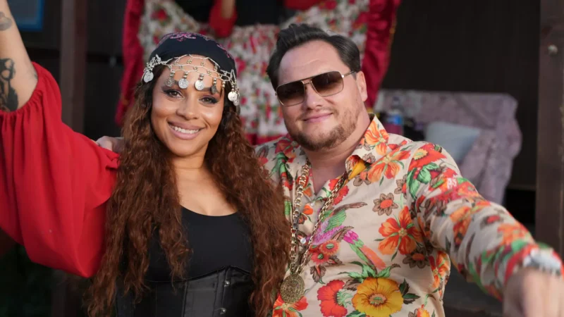 Na fotografii sú dvaja ľudia v pestrofarebných oblečeniach s úsmevmi na tvári. Žena má dlhé vlnité vlasy, červenú blúzu a ozdobnú čelenku s mincami, muž má kvetovanú košeľu, zlatý reťaz a slnečné okuliare. Obaja stoja blízko seba a pôsobia, akoby sa zabávali na tematickej oslave alebo festivale.