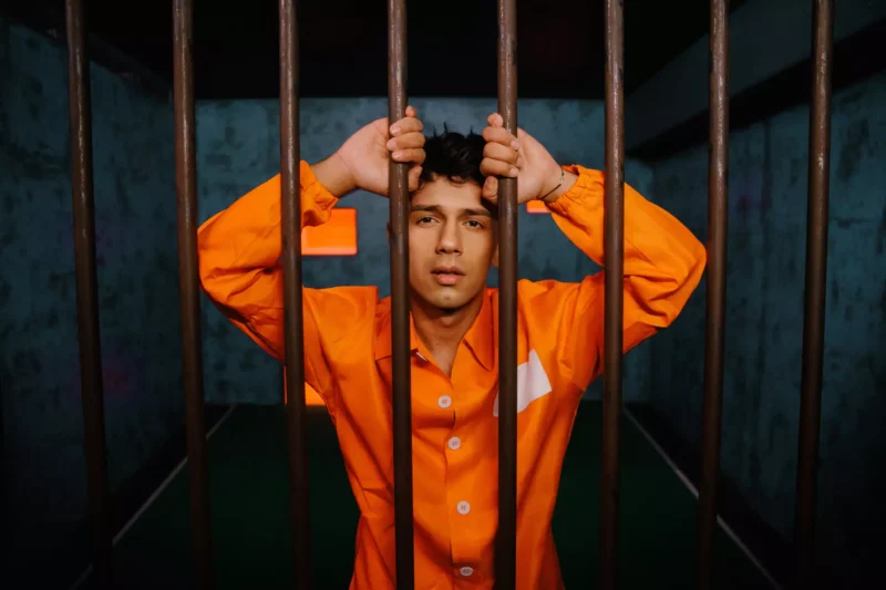 Muž v oranžovej väzenskej kombinéze sa drží kovových mriežok vo vnútri cely a vyzerá zachmúrene. Tlmené svetlo a modré steny vytvárajú napätú atmosféru.